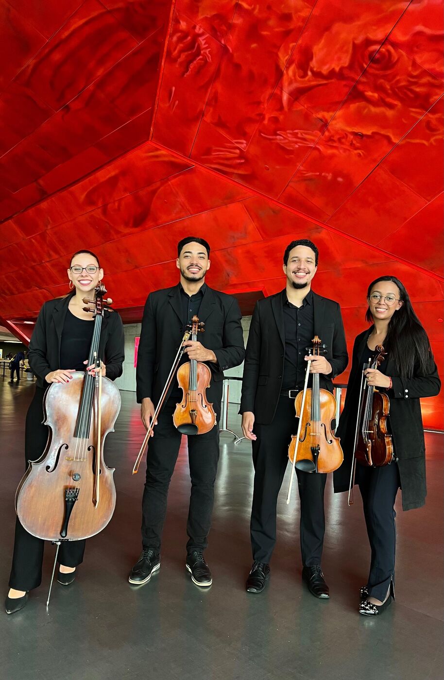 Quarteto de cordas posando com os instrumentos diante de uma parede vermelha