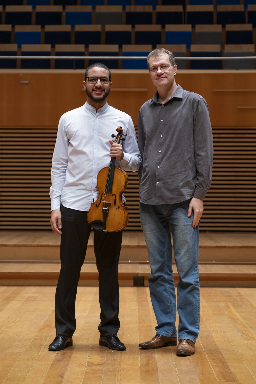 André com a viola no palco de uma sala de concertos, ao lado de um professor